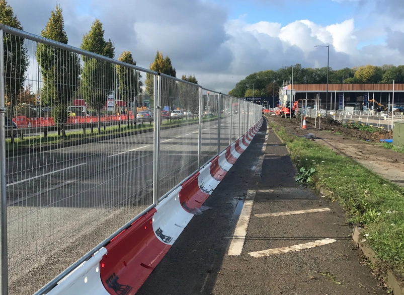 Temporary roadworks safety fencing and red and white bull barriers along a roadside footpath